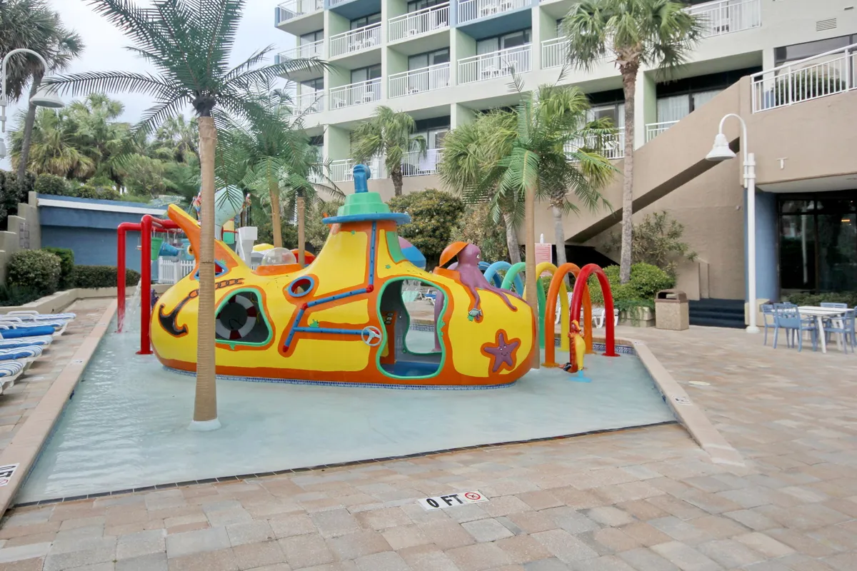 The photo features a colorful submarine-themed water play structure for children, surrounded by palm trees and lounge chairs. Kids are seen playing and laughing in the shallow splash zone, making it an energetic, family-friendly scene.