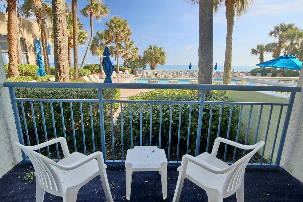 A serene exterior scene showcasing balcony seating overlooking a sparkling pool area, with swaying palm trees and the ocean visible in the distance. The inviting space hints at opportunities for relaxation, conversation, and enjoying coastal sunsets, all set in a lush, tropical environment.