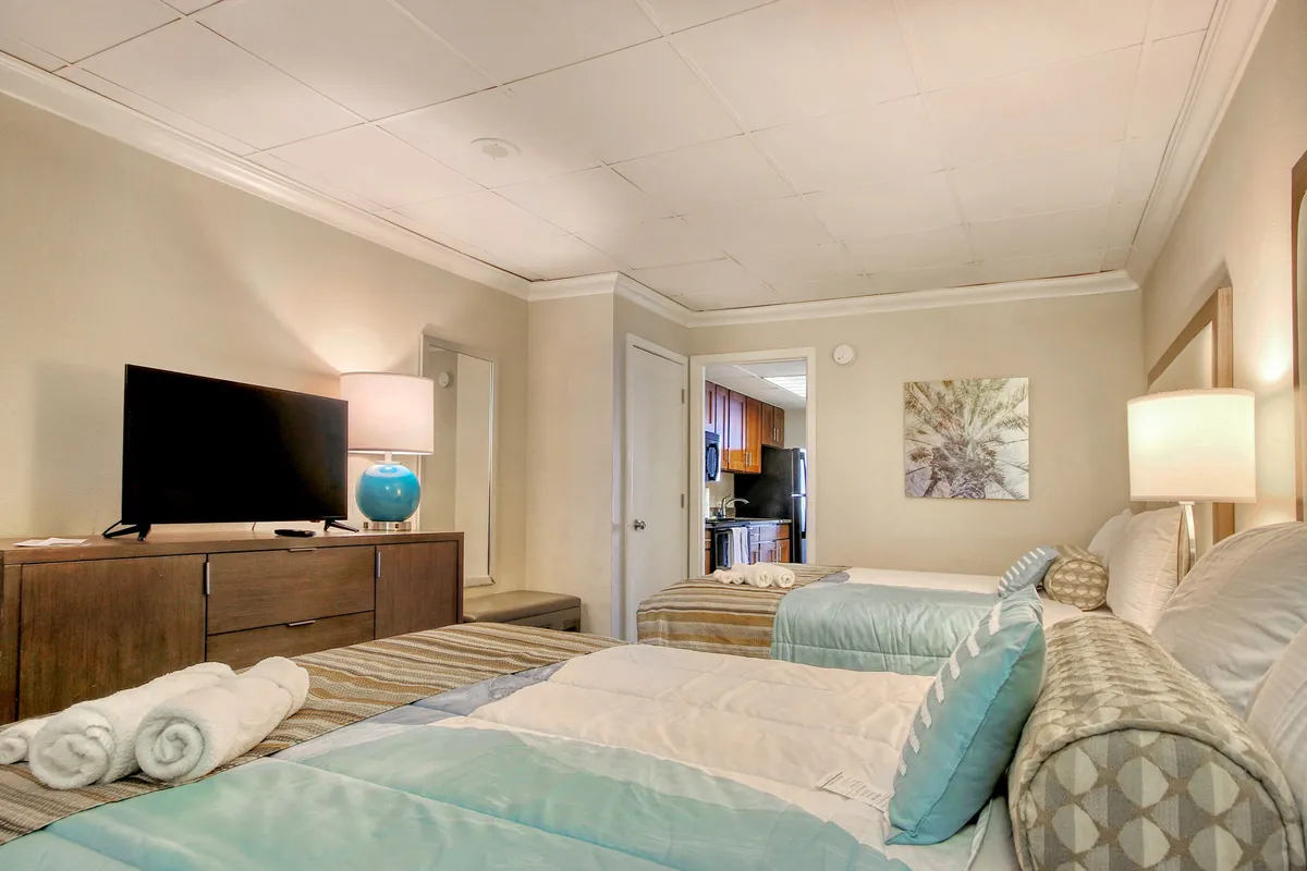 A cozy bedroom setup with two beds adorned with decorative pillows, a television unit with a lamp, and a piece of calm artwork above. The layout encourages relaxation, whether you're unwinding with a show or sharing quiet moments with family.