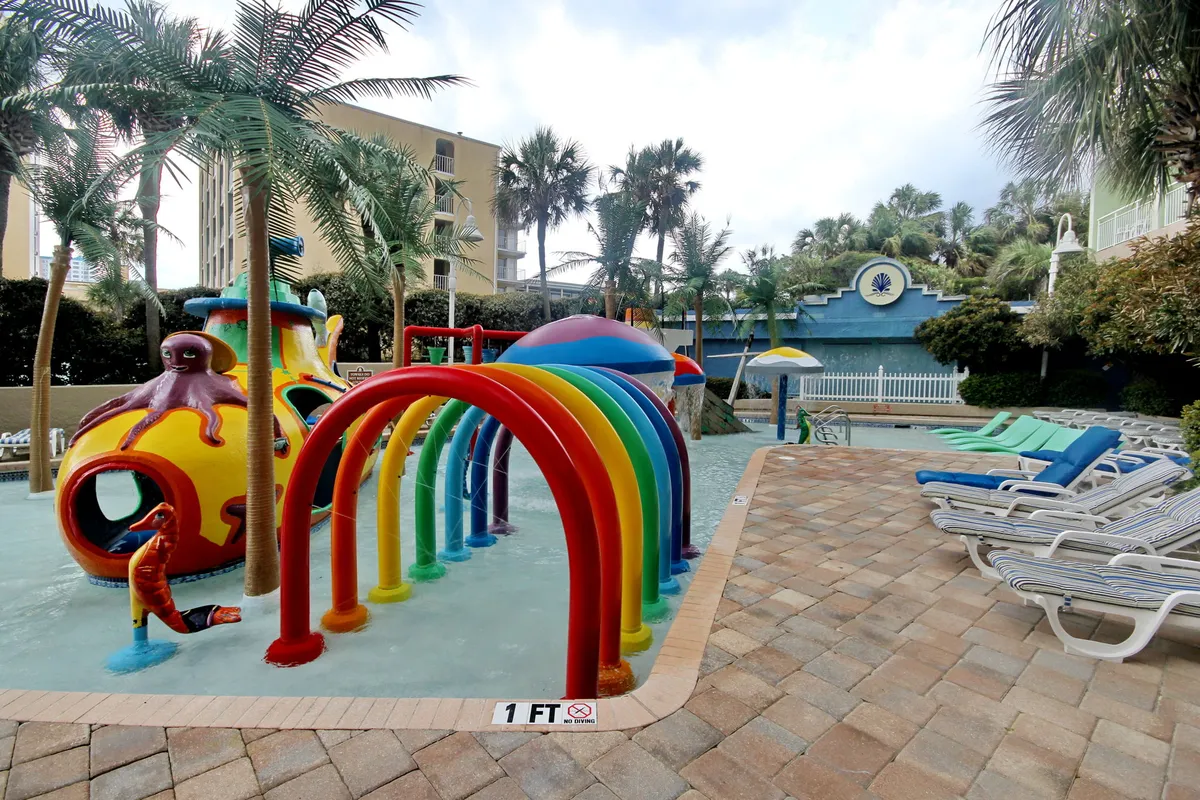 A vibrant outdoor pool area featuring a colorful water play structure, rainbow arches, and poolside lounge chairs. Children are seen playing joyfully, with families gathered around and adults relaxing nearby. The entire space is bathed in sunlight, emphasizing fun and lively activity.