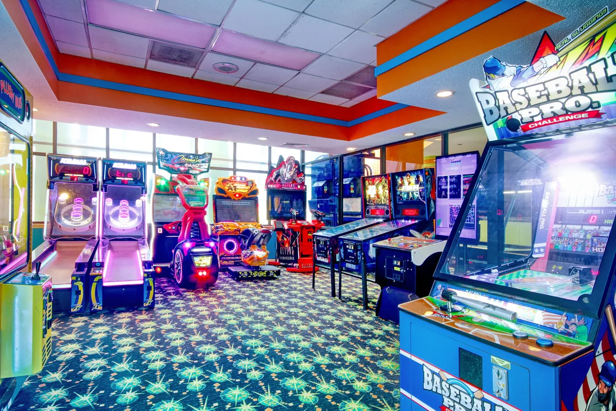 An interior shot of the resort’s arcade area, showing rows of classic arcade machines, a brightly colored carpet, and ample natural light from large windows. Families and groups enjoy games together, fostering a sense of nostalgia and fun.