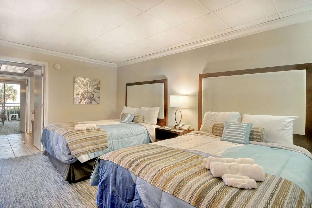 The image features a serene interior scene with two inviting beds adorned with decorative pillows, a softly glowing nightstand lamp, and tasteful coastal artwork on the wall. Gentle natural light filters through, creating a bright and peaceful atmosphere perfect for unwinding.