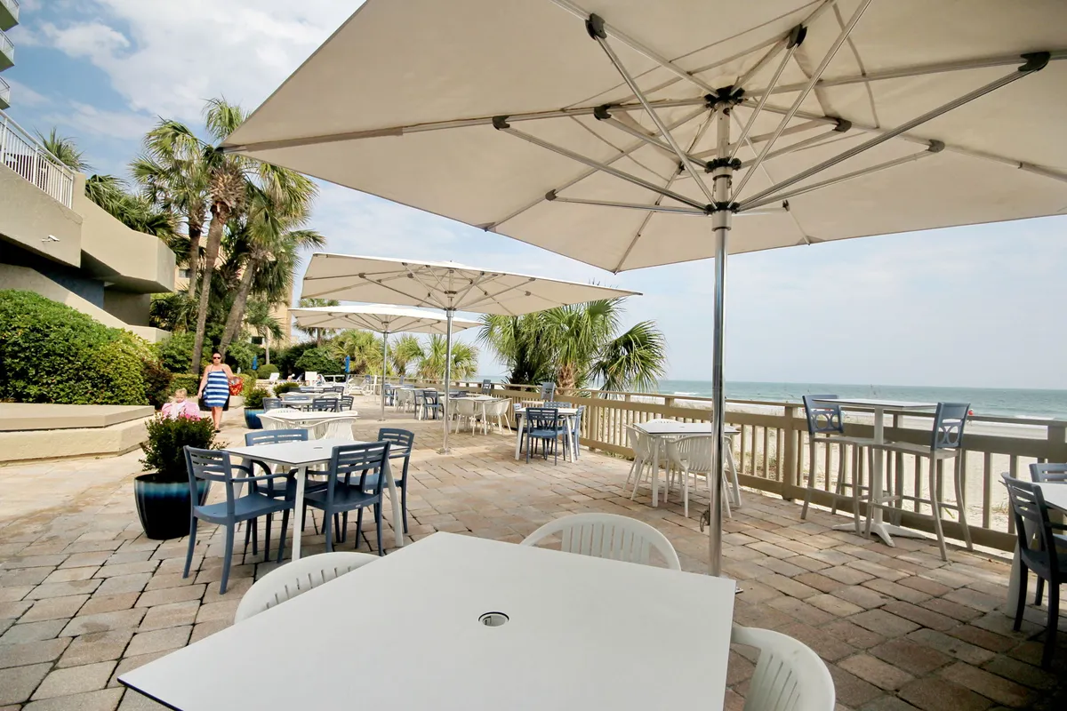 An inviting outdoor dining area with tables shaded by umbrellas, surrounded by palm trees and facing a breathtaking ocean view. The casual setting suggests opportunities to enjoy al fresco meals, tropical drinks, and long conversations while taking in the beauty of the coast.