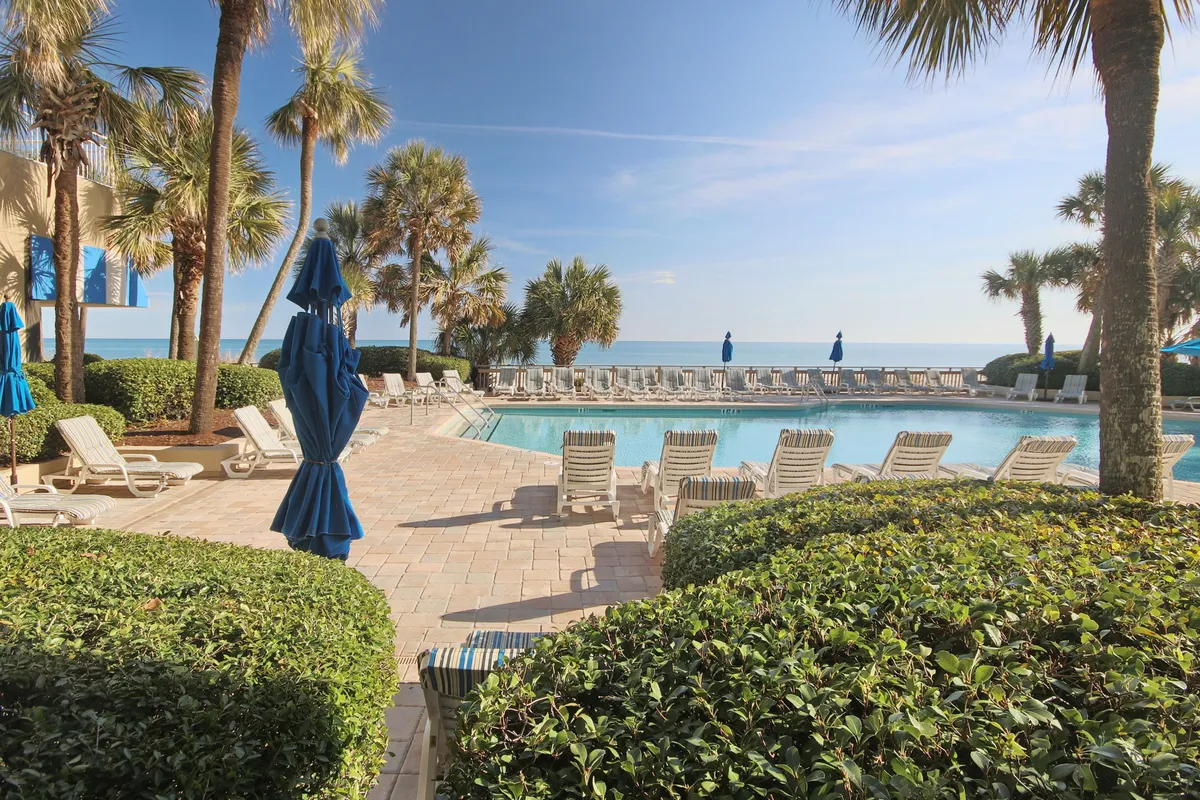 This exterior image captures a sparkling swimming pool surrounded by inviting lounge chairs and swaying palm trees. Blue umbrellas pop against the tranquil backdrop, and the ocean is visible beyond the pool area, enhancing the resort's vacation atmosphere.