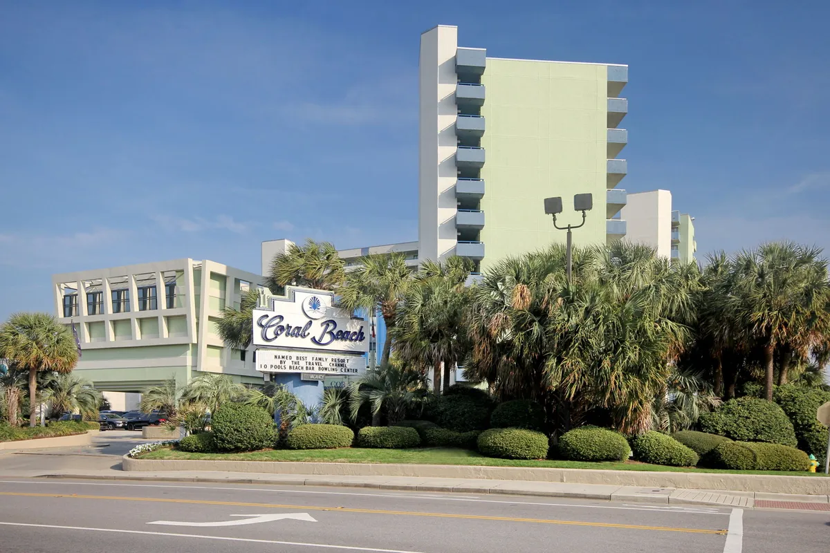 A wide shot of the Coral Beach Resort exterior, featuring the prominent Coral Beach sign, green building, and lush palm trees. The image conveys immediate beach and vacation vibes, with the resort itself as the main subject. Hints of the pool and proximity to the beach are visible, suggesting both direct access to the shoreline and the range of resort amenities available.